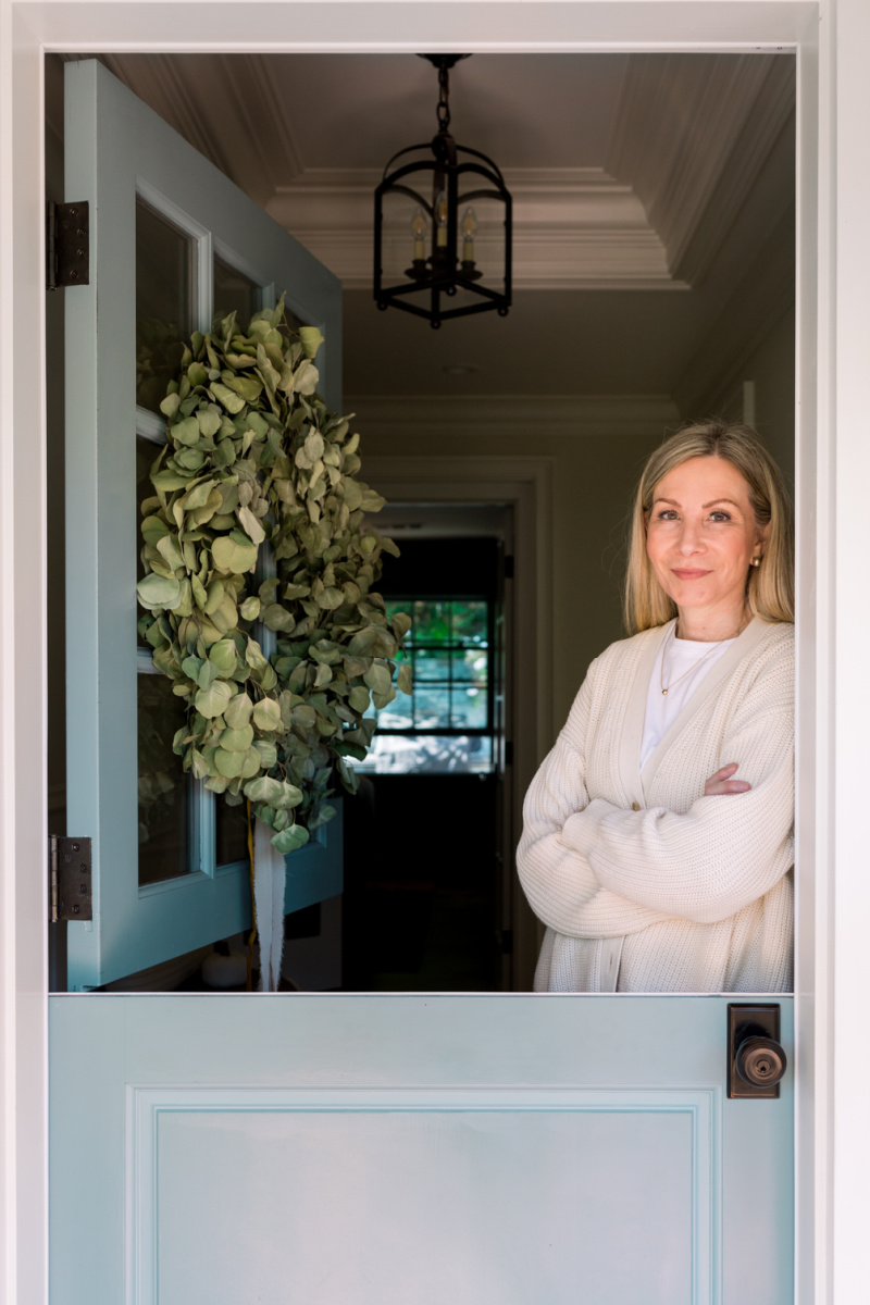 Woman standing inside blue Dutch door with eucalyptus wreath