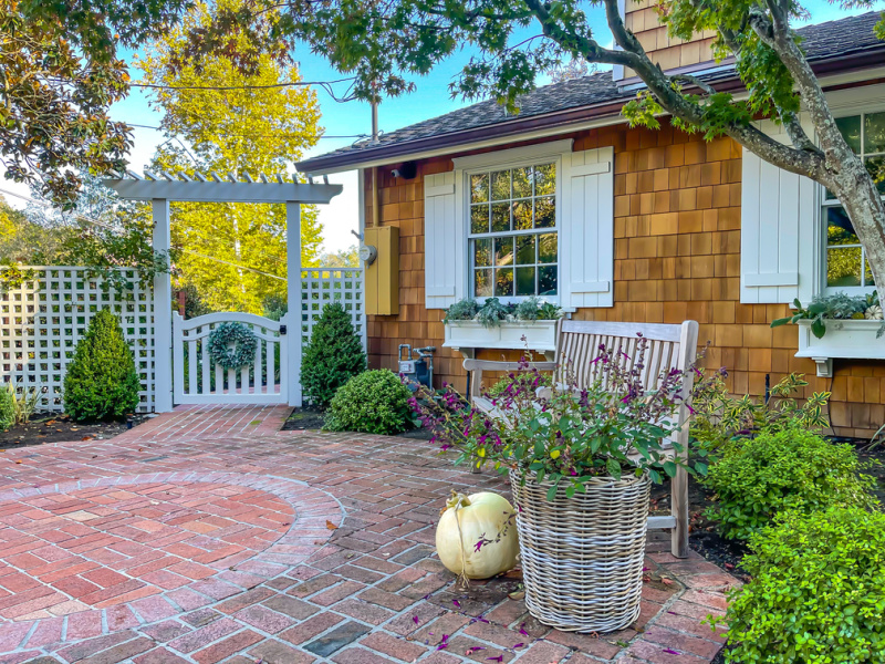 Front entry of shingled house with shutters and window boxes and white pumpkin next to planter and bench.