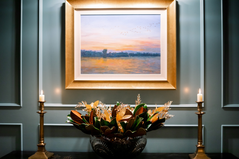 Punch bowl filled with magnolia stems flanked by brass candlesticks and art.