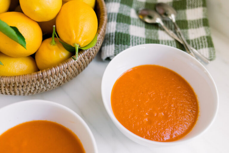 Bowls of tomato soup next to a bowl of lemons and green and white dishcloth.