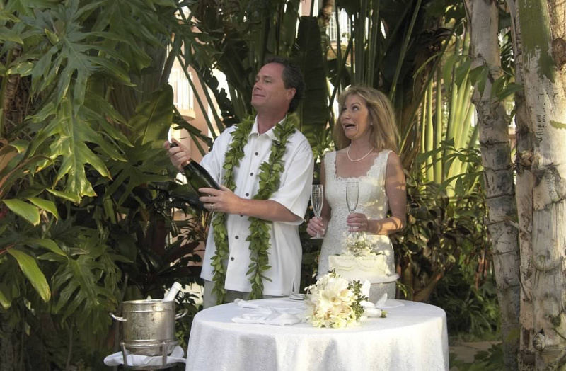Bride and groom in front of wedding cake popping cork from champagne bottle.