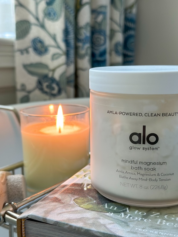 Alo Bath Soak jar on bath tub tray next to burning candle.