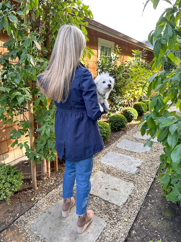 Woman wearing Boden utility jacket and holding little white dog in garden.