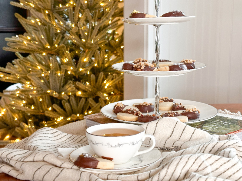 Tiered tray filled with holiday cookies on table next to cuppa tea with Christmas tree in background.