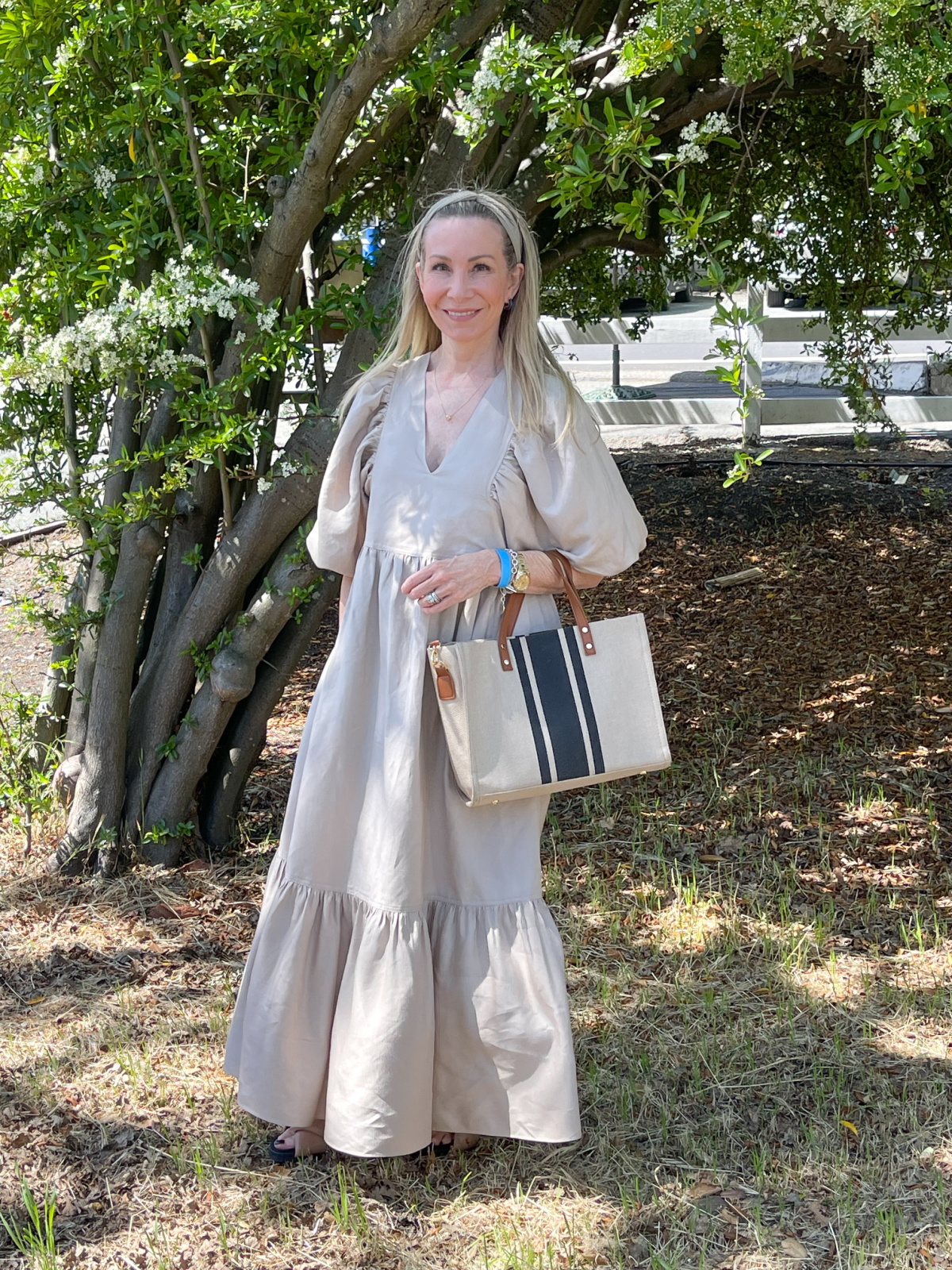 Woman wearing beige dress holding striped tote bag standing under tree.