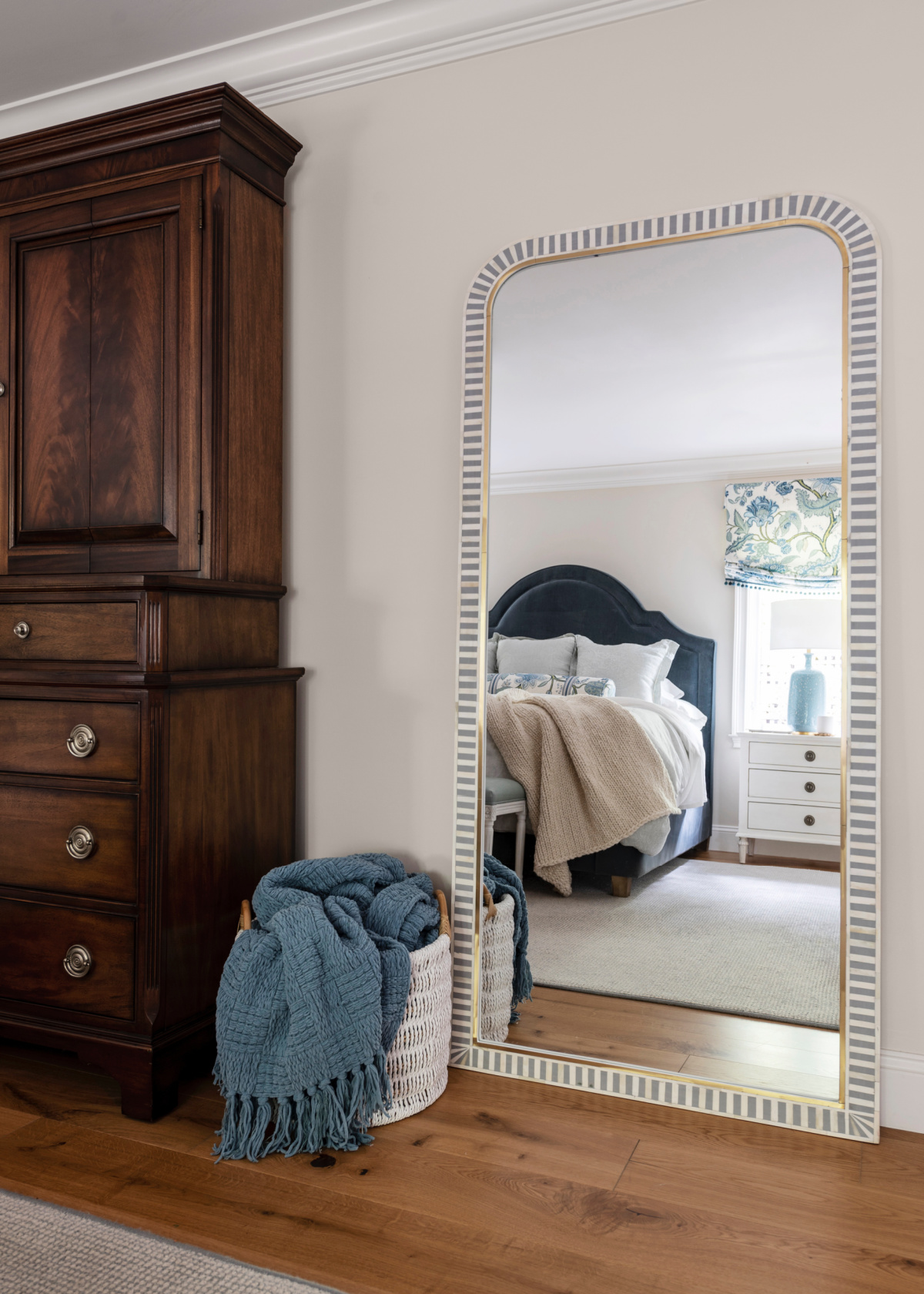 Bedroom floor mirror next to armoire reflecting the bed on other side of the room.