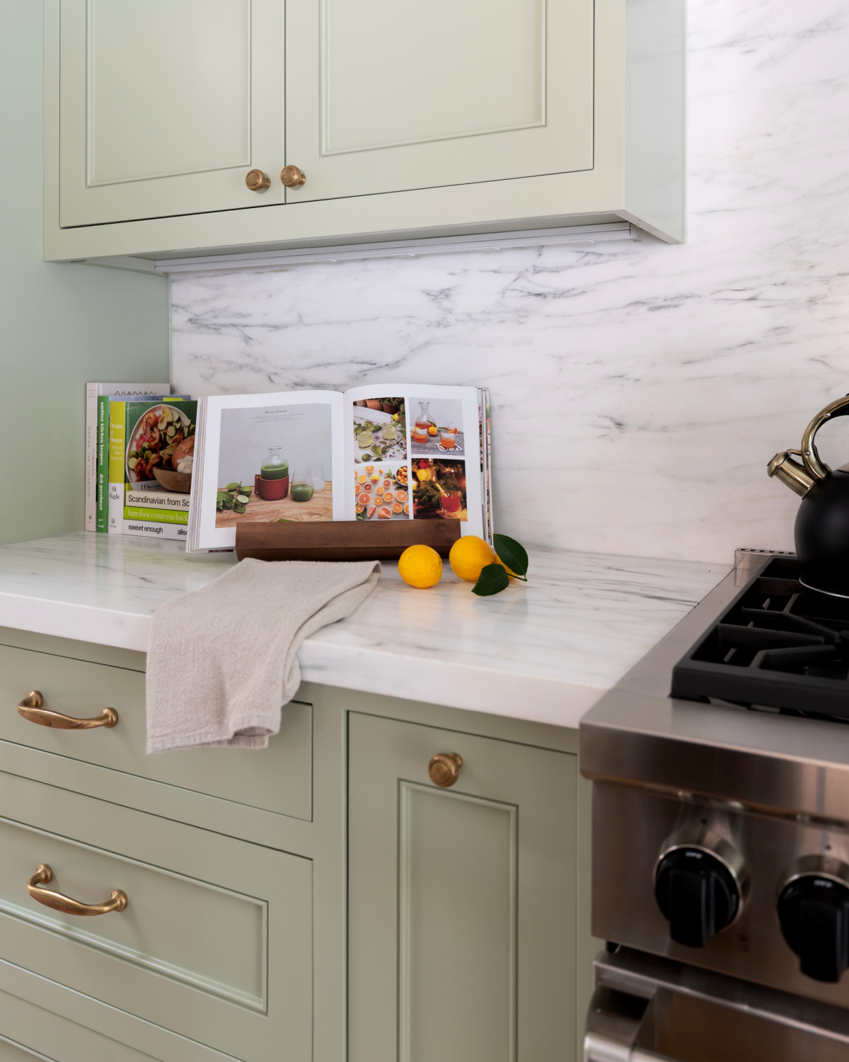 Kitchen counter vignette with standing cookbooks and open cookbook on cookbook stand next to lemons and a tea towel.
