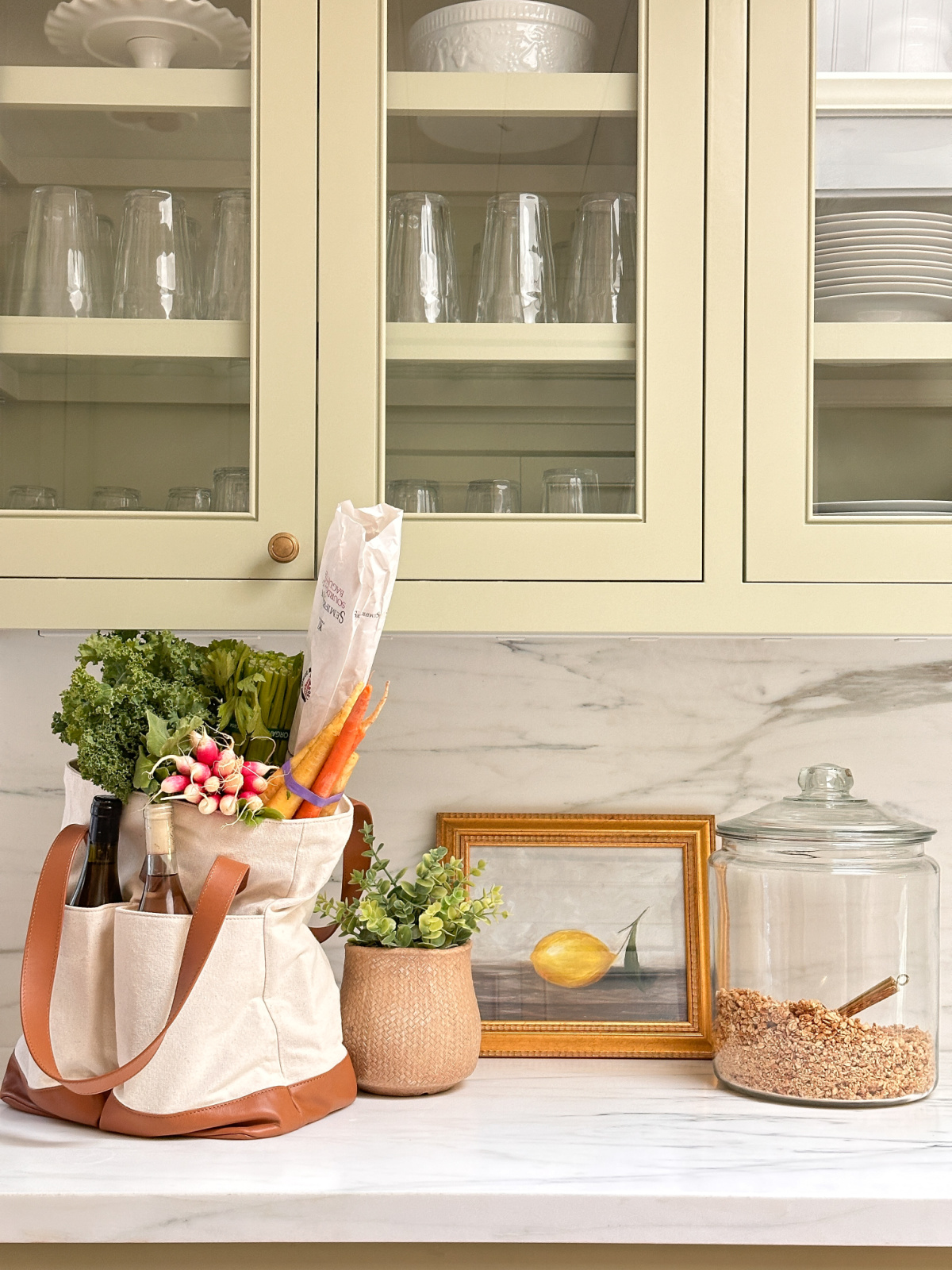Canvas and leather tote bag filled with produce and groceries on kitchen counter next to plant, lemon artwork and a jar full of granola.