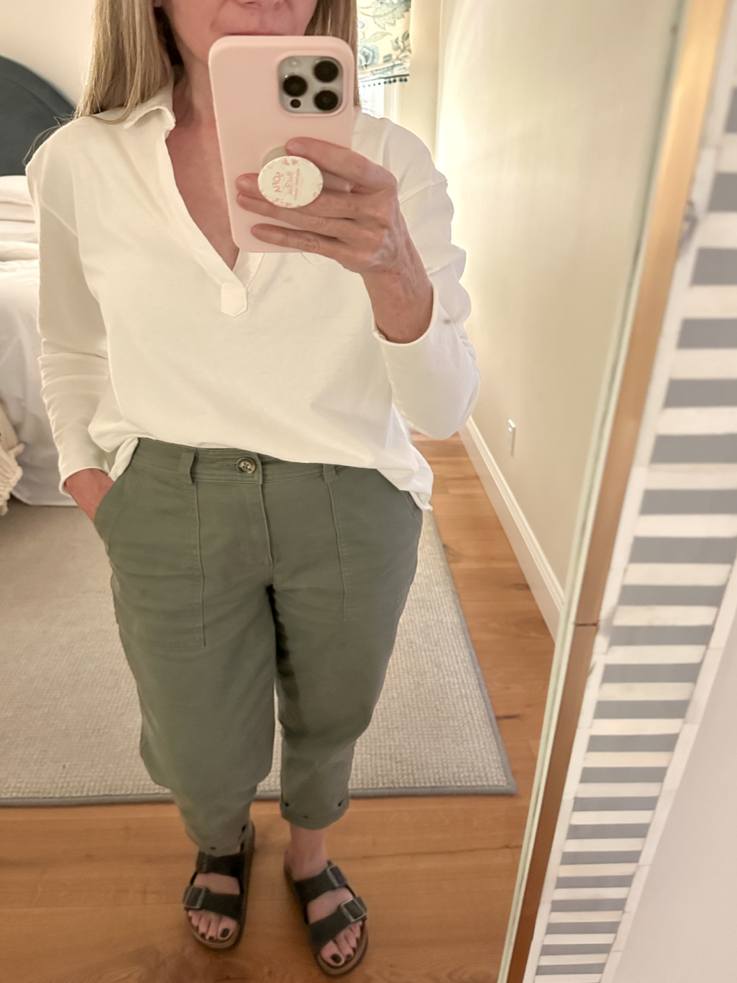 Woman taking mirror selfie wearing white shirt, green pants and Birkenstocks.