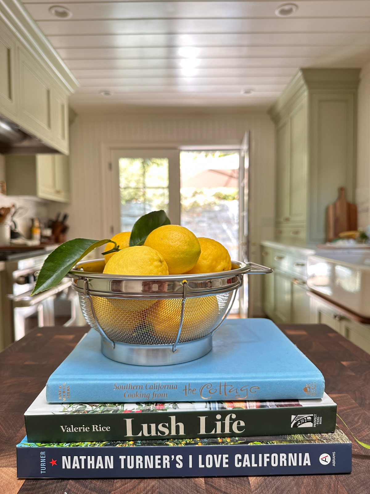 Collander of lemons sitting on stack of three books on kitchen island with green galley kitchen in background.