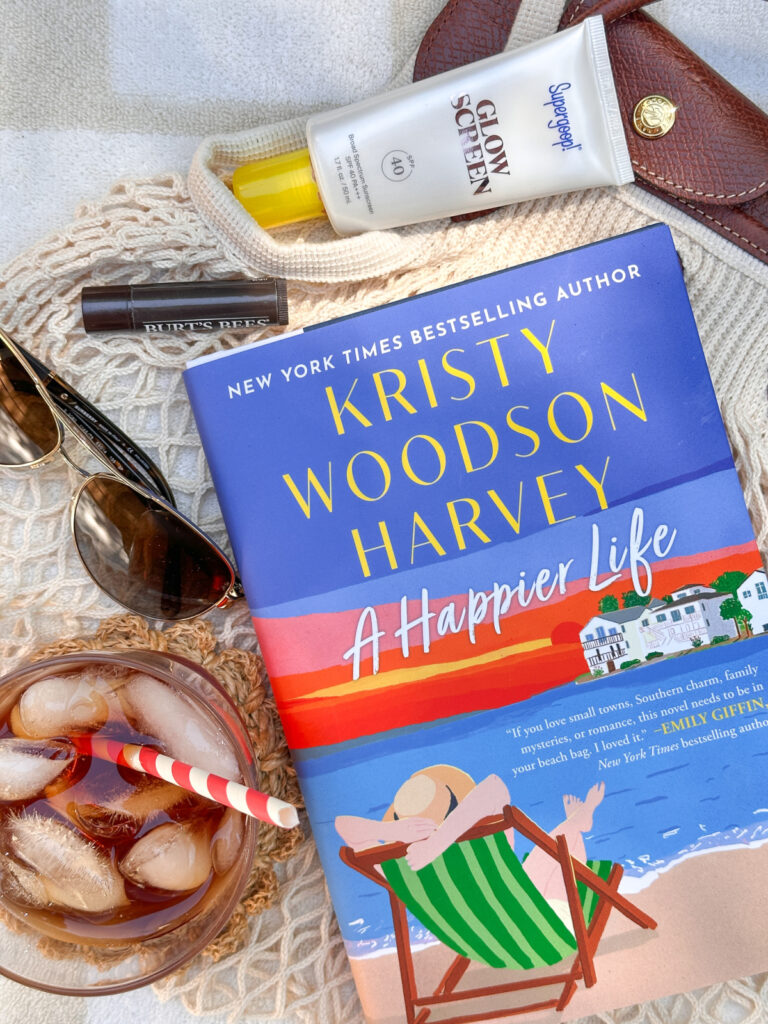 A Happier Life book on beach towel next to beach bag, sunglasses, sunscreen and a cold drink.