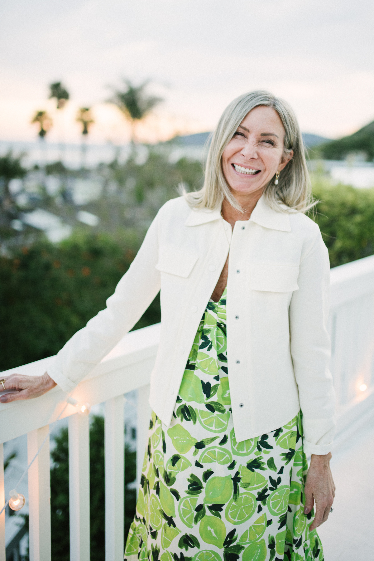 Woman standing on ocean front balcony wearing lime print dress and ivory jacket.