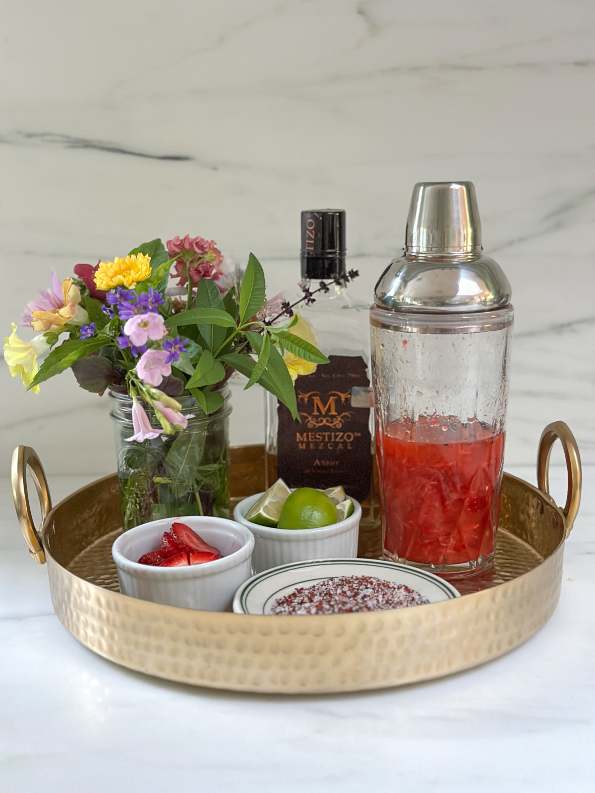 Copper tray with a vase of flowers, mezcal, cocktail shaker and ingredients.