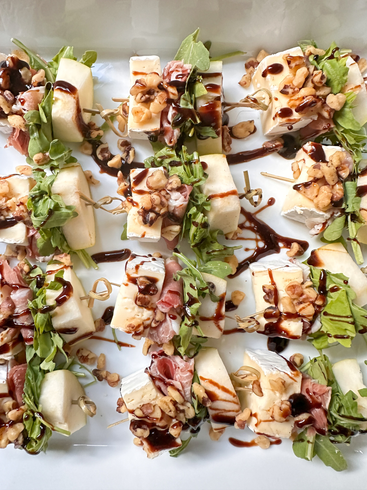 Pear, brie, prosciutto and arugula skewers on a white platter.