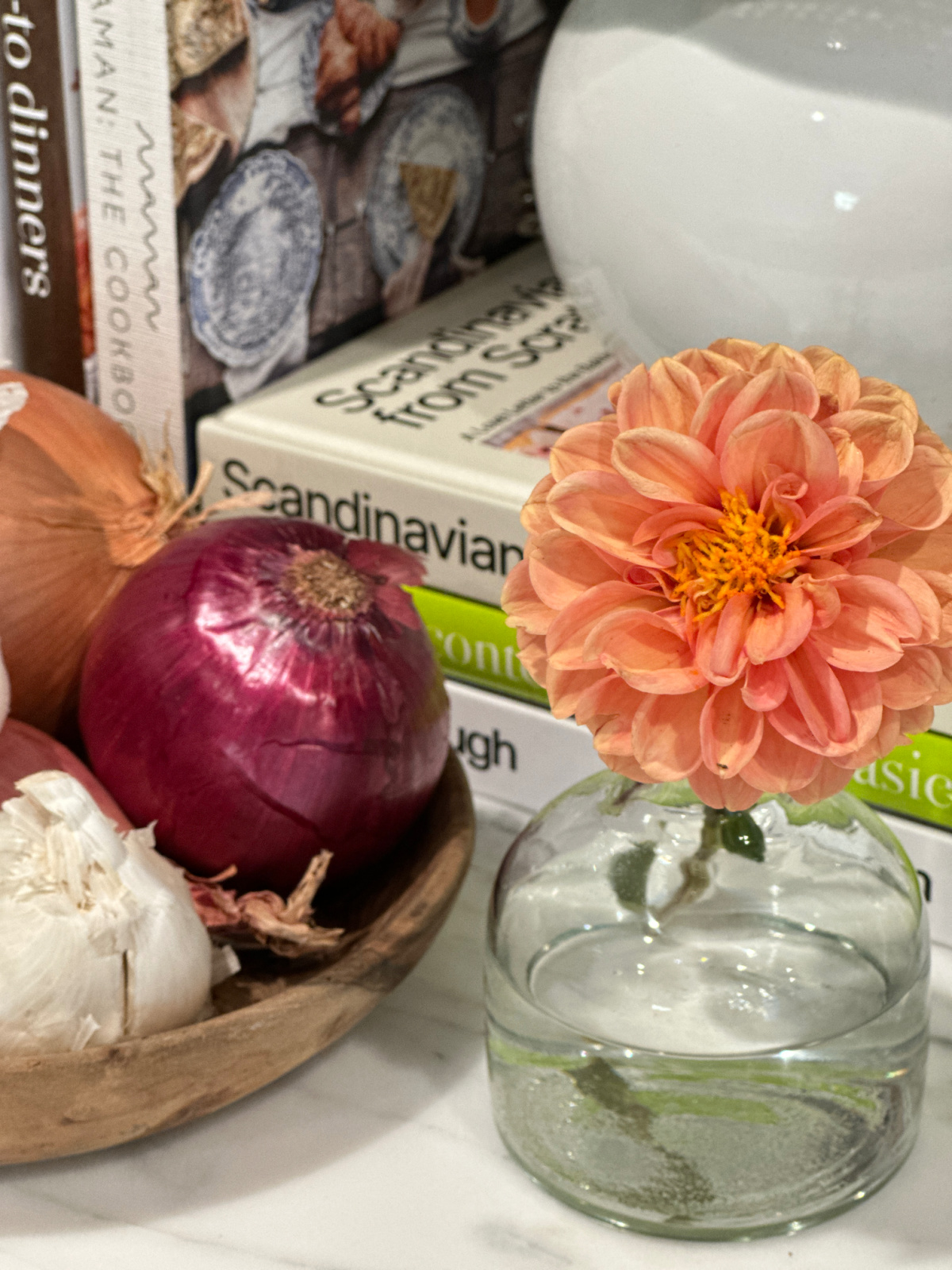Orange dahlia in bud base next to wooden bowl of onions and garlic.