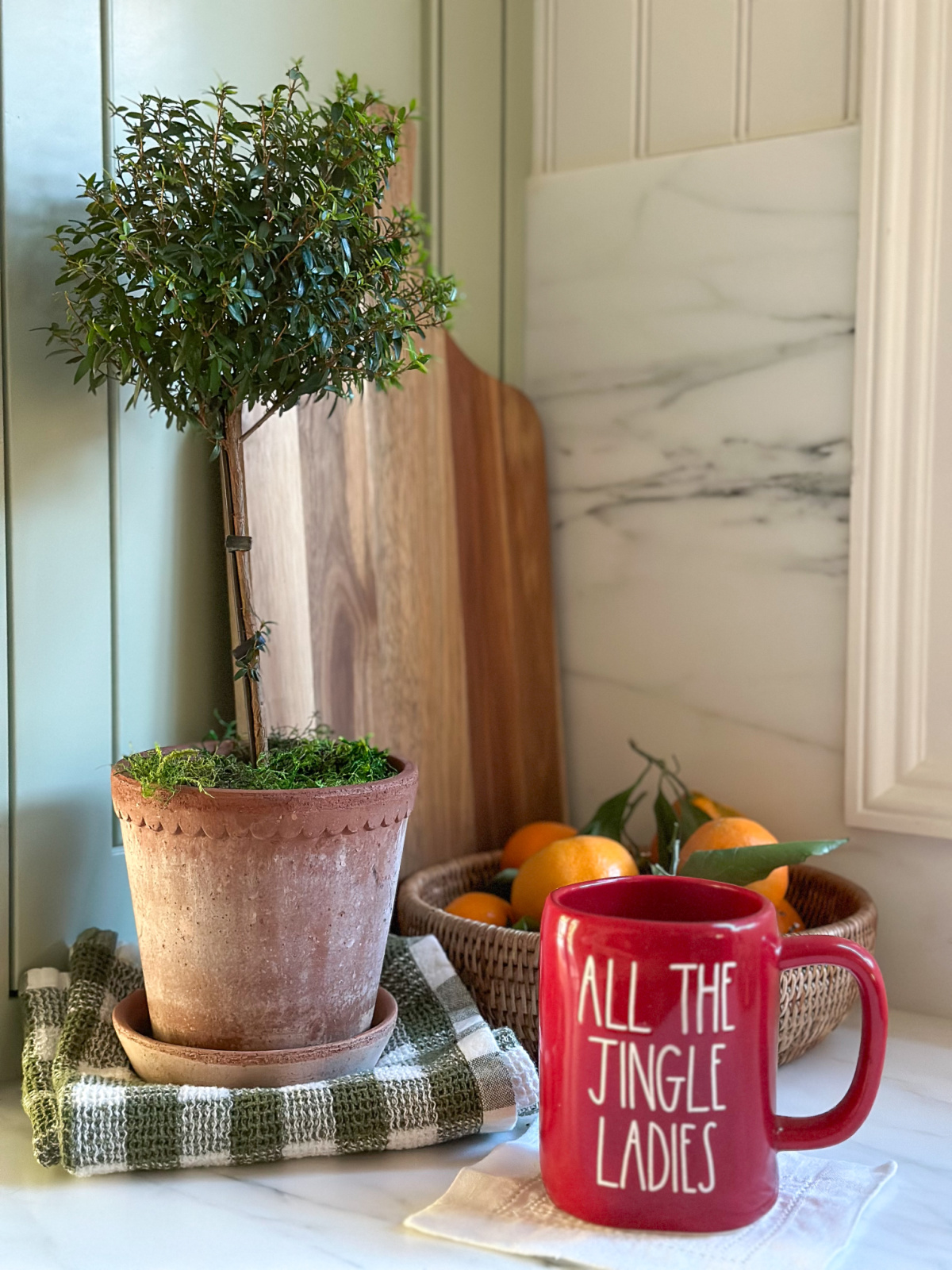 Christmas mug on counter next to bowl of oranges and topiary.