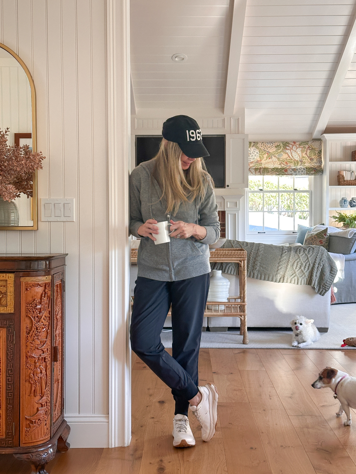 Woman wearing cashmere hoodie, holding mug of cocoa, looking at little dogs.