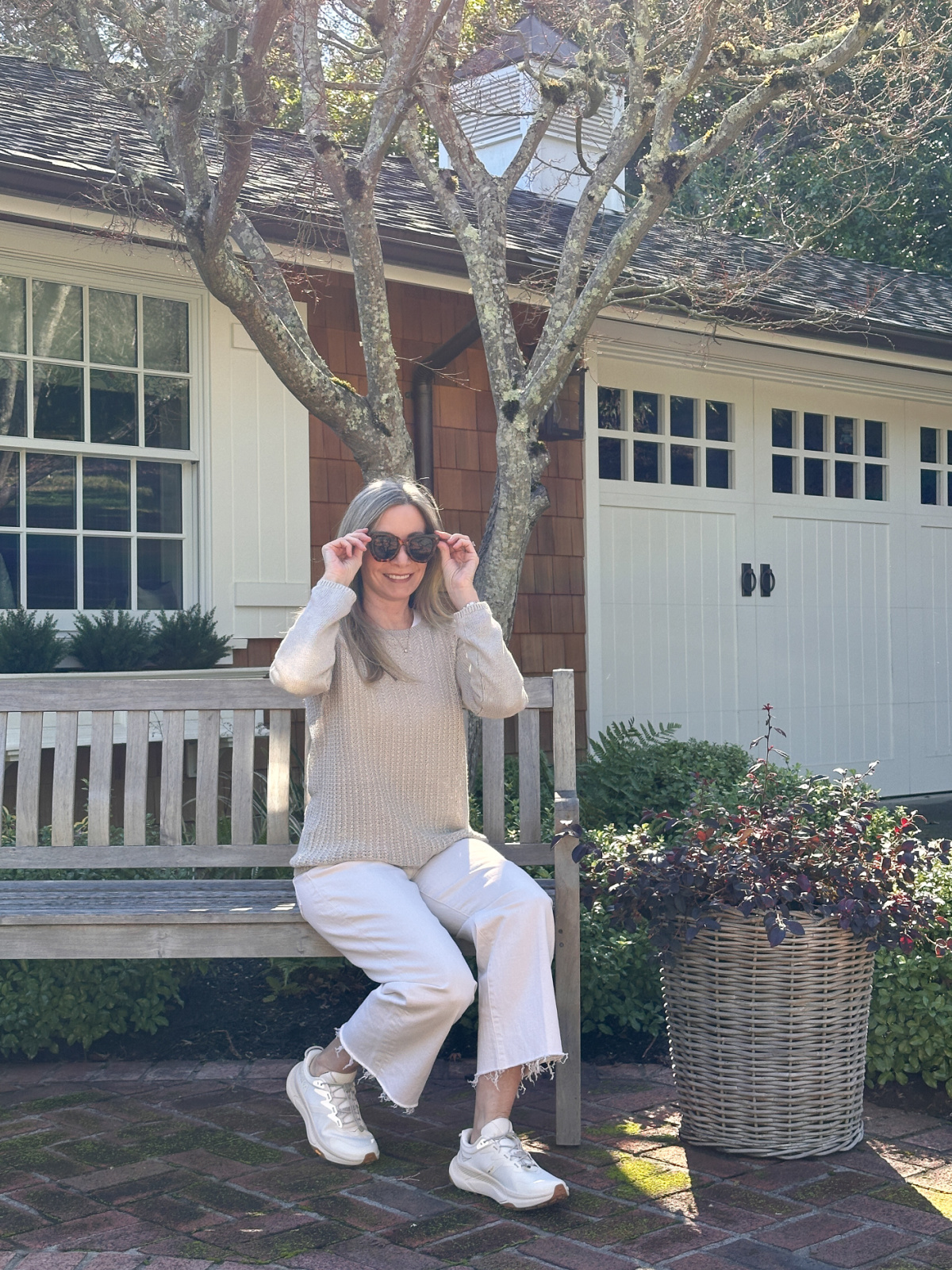 Woman wearing white denim, beige sweater and sneakers sitting on bench.