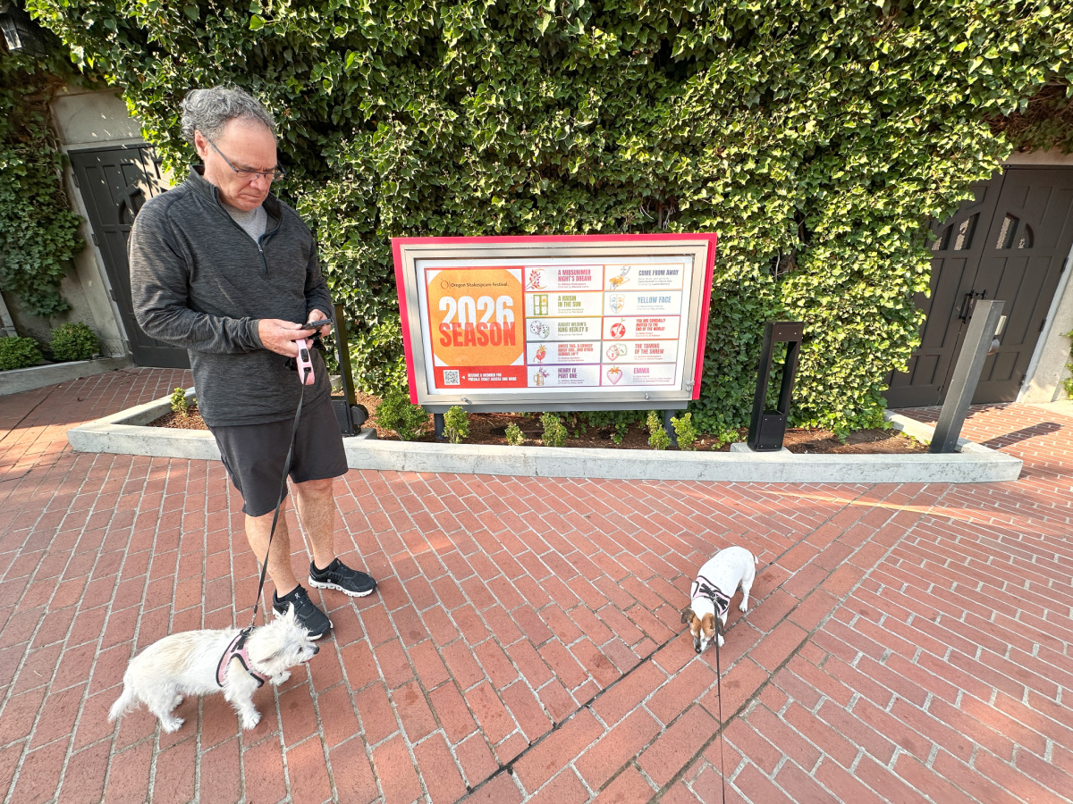 Man and two small dogs in front of Oregon Shakespeare Festival 2026 season announcement.